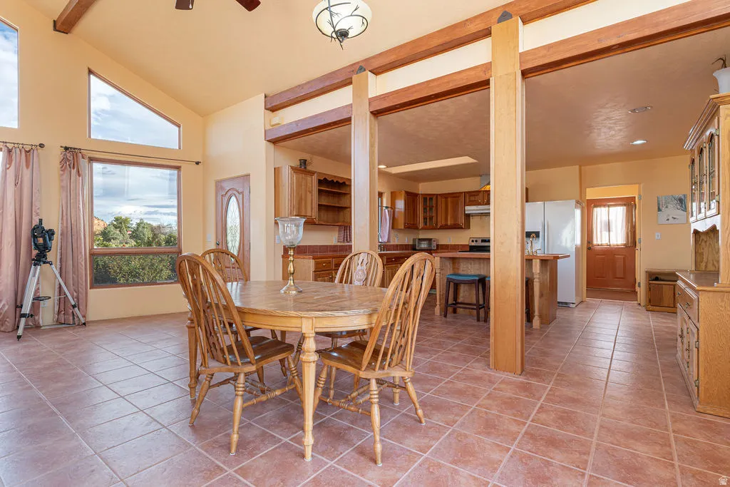 Spacious open-concept dining and kitchen area featuring soaring ceilings with exposed wood beams, large windows with natural light, terra cotta tile flooring, and substantial wooden support columns defining the layout