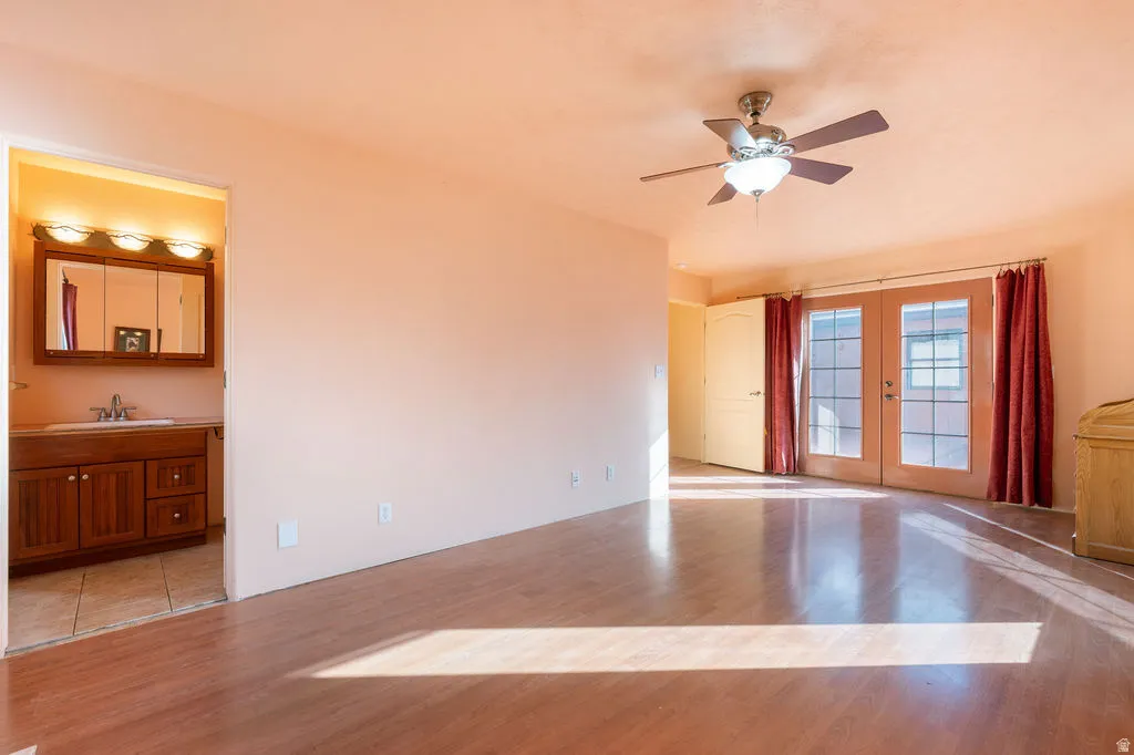 Wood-finish flooring throughout, ceiling fan with integrated lighting, glass-paneled French doors, and an attached bathroom featuring a wood vanity with integrated sink and triple vanity lighting