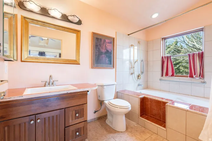 Bathroom featuring a wood-finish vanity with a single basin sink, gold-framed mirror, and a tiled bathtub surround with an integrated shelf