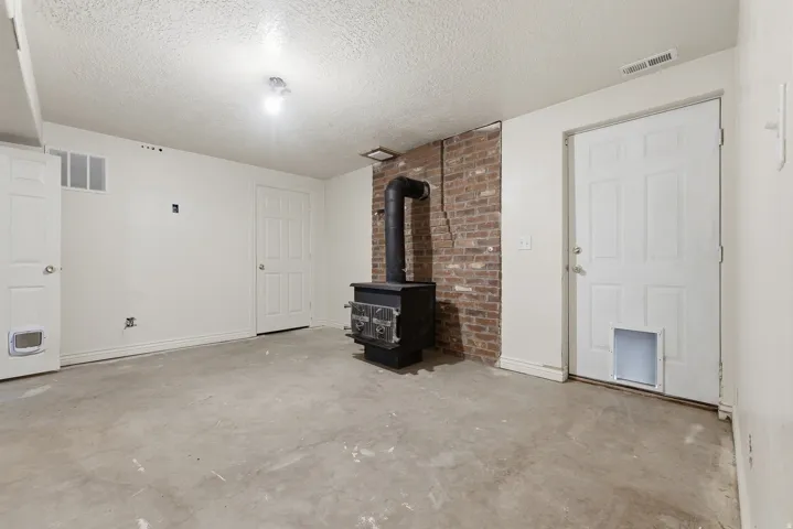 Spacious basement featuring exposed brick accent wall, concrete flooring, wood-burning stove with chimney, and multiple white finished doors providing access to adjoining spaces. Exterior entrance door