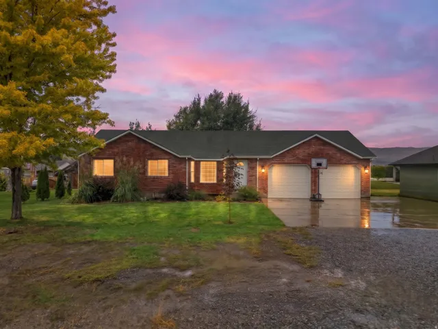 Brick ranch home with illuminated interior, featuring a two-car garage, manicured lawn with mature landscaping, and scenic mountain backdrop at dusk