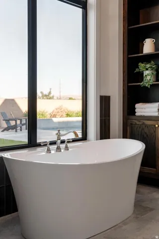 Freestanding soaking tub with polished chrome fixtures, full-height window, dark wood-finish built-in shelving, and dark floor tiling