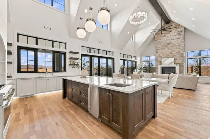 Vaulted ceiling with exposed beam, stone-clad fireplace, wood-finish flooring, kitchen island with built-in sink, and integrated stainless steel dishwasher