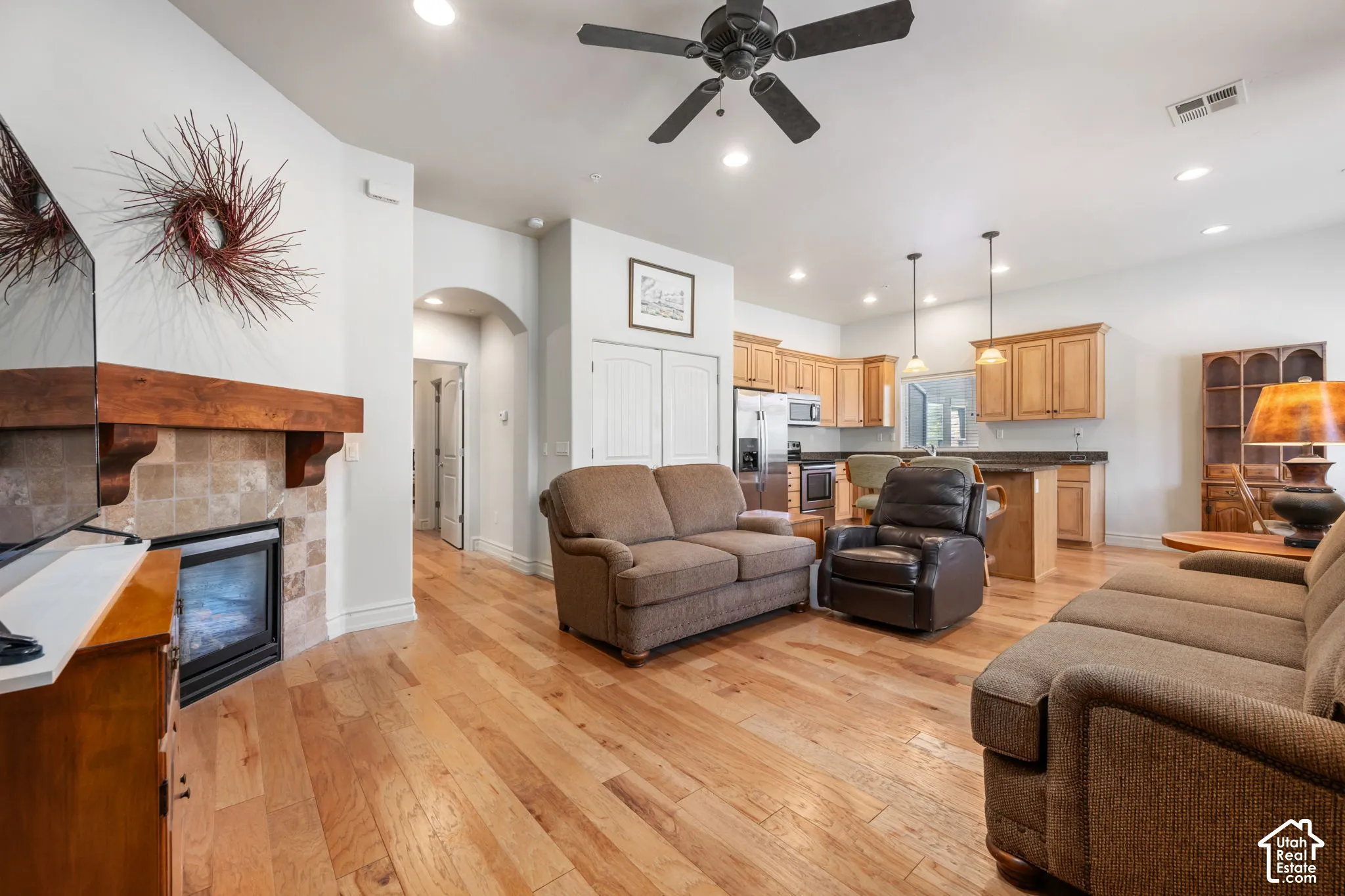 Wood-finish flooring throughout, living area with fireplace featuring a wood mantel and tile surround, kitchen with wood cabinetry, stainless steel appliances, and a breakfast bar, and recessed lighting
