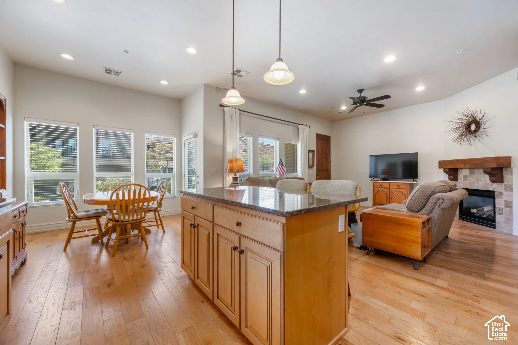 Kitchen island with a dark countertop and light wood-finish cabinetry, wood-finish flooring, recessed lighting, and a fireplace with a tile surround and wood mantel