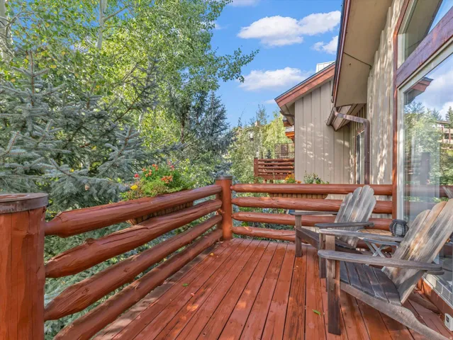 Expansive wood-finish deck with a log railing, featuring surrounding mature trees and a large glass door