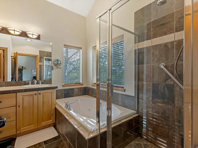 Vaulted ceiling, wood-finish vanity, integrated soaking tub, separate glass-enclosed shower with tile surround, and tile flooring