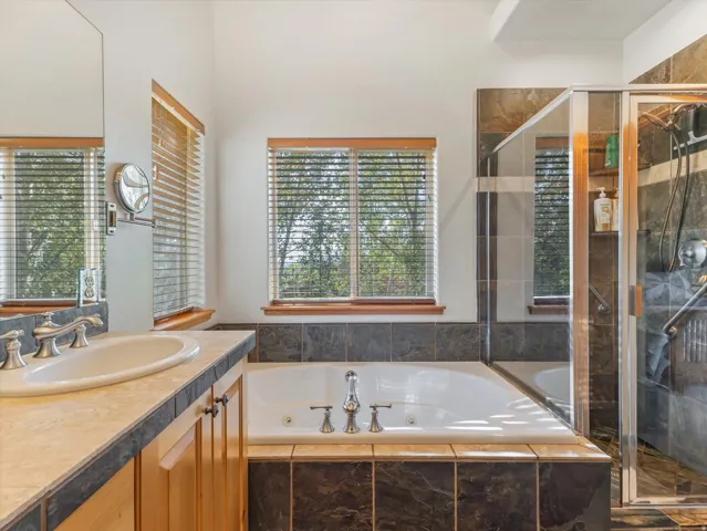 Built-in jetted tub, glass-enclosed shower, wood-finish vanity with an integrated sink, tiled countertop, and large windows with blinds