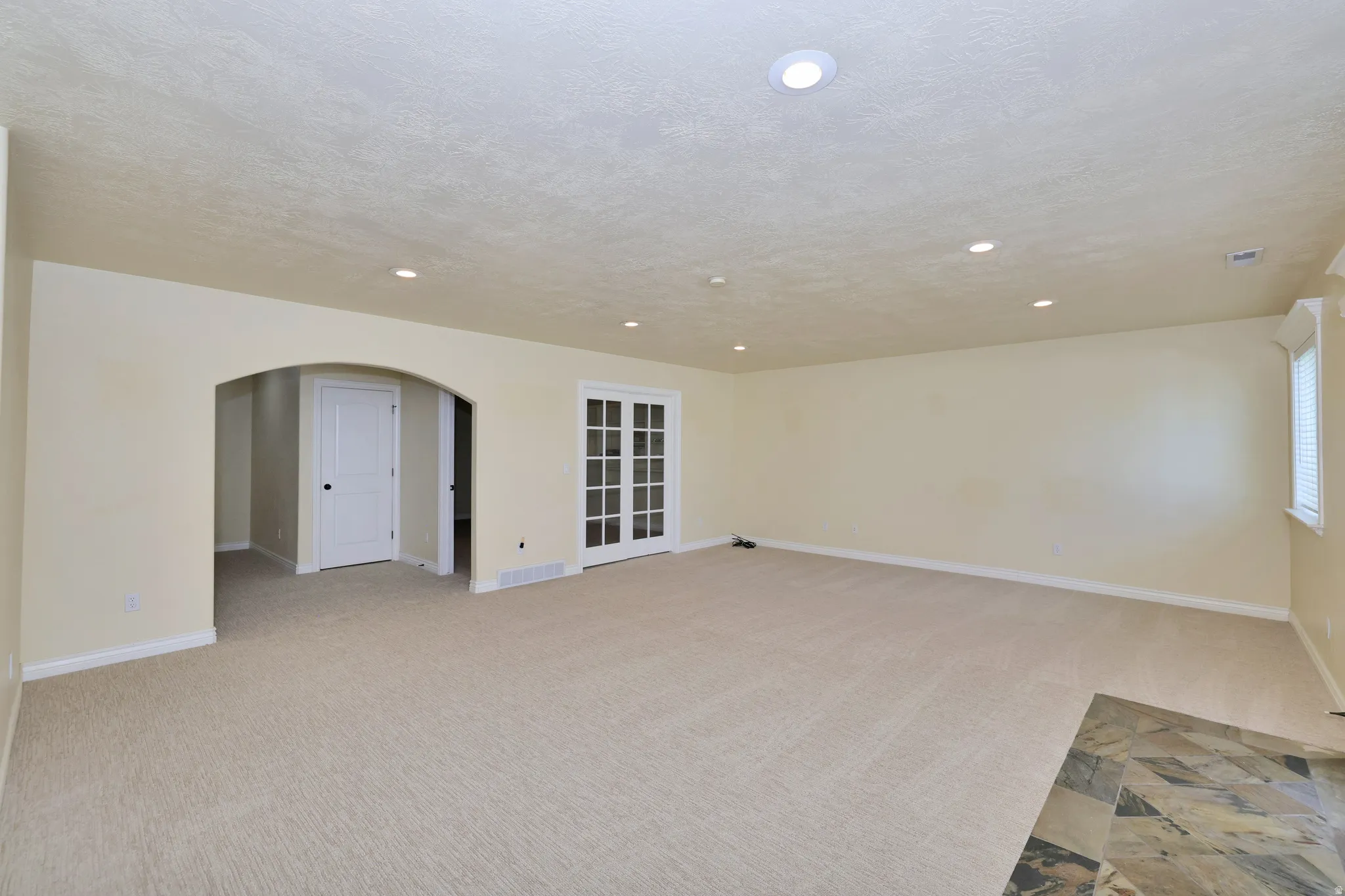 Recessed lighting illuminates the carpeted room, featuring an arched doorway, French doors, and a tiled floor section