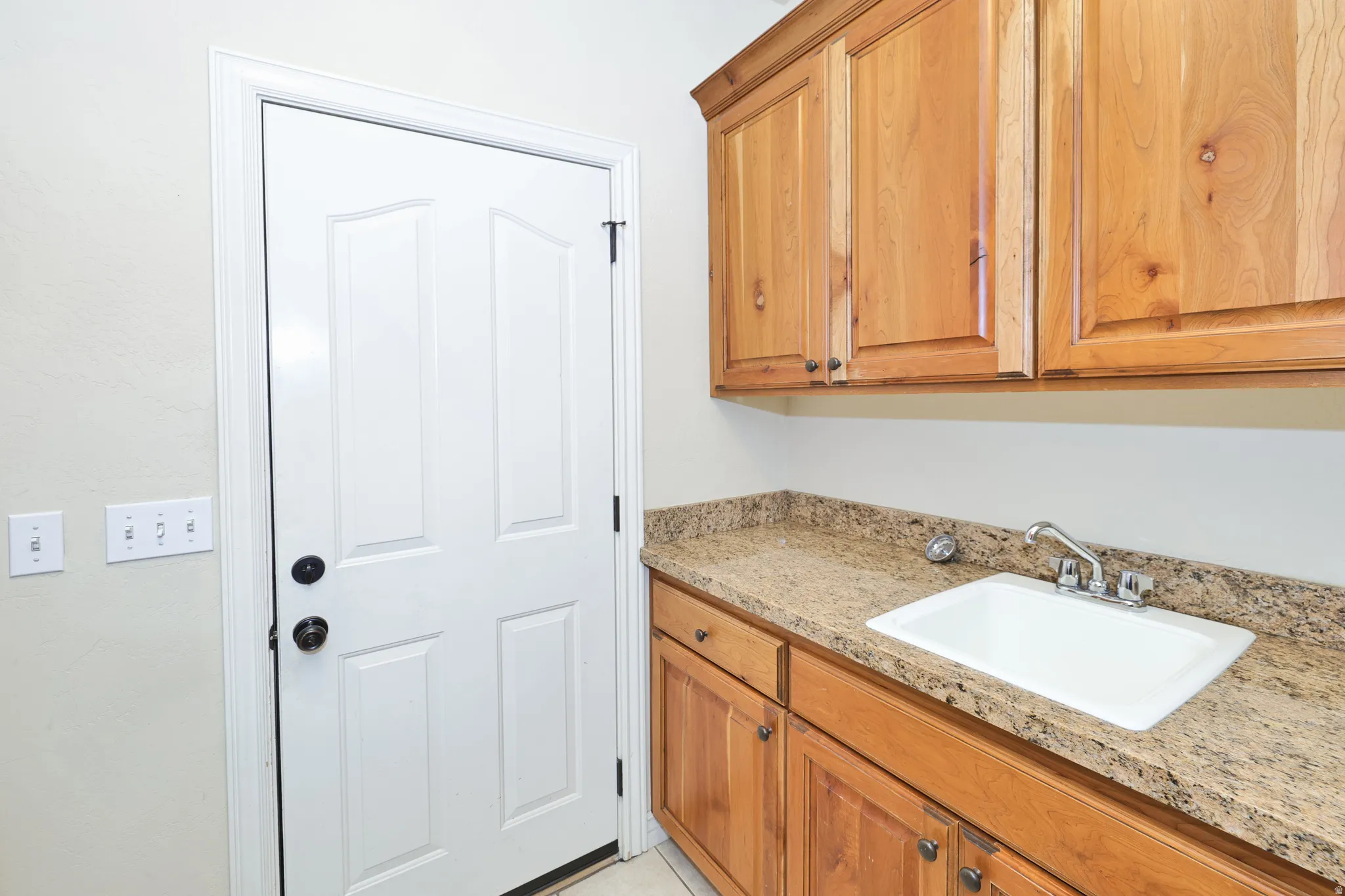 Utility room featuring natural wood cabinetry with panel doors, granite-style countertop with undermount sink, and brushed nickel faucet