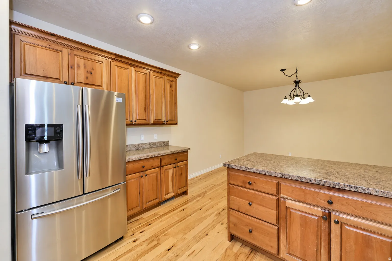 Stainless steel French door refrigerator, wood-finish flooring, recessed lighting, wood cabinetry, and a central island with a stone-finish countertop