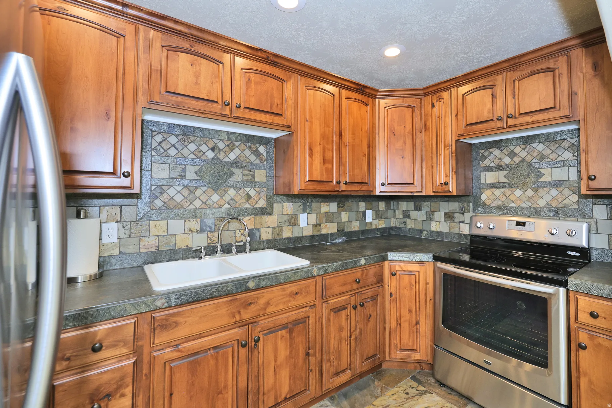 Wood cabinetry, double basin sink, stone-finish countertops, stainless steel range, and tile backsplash