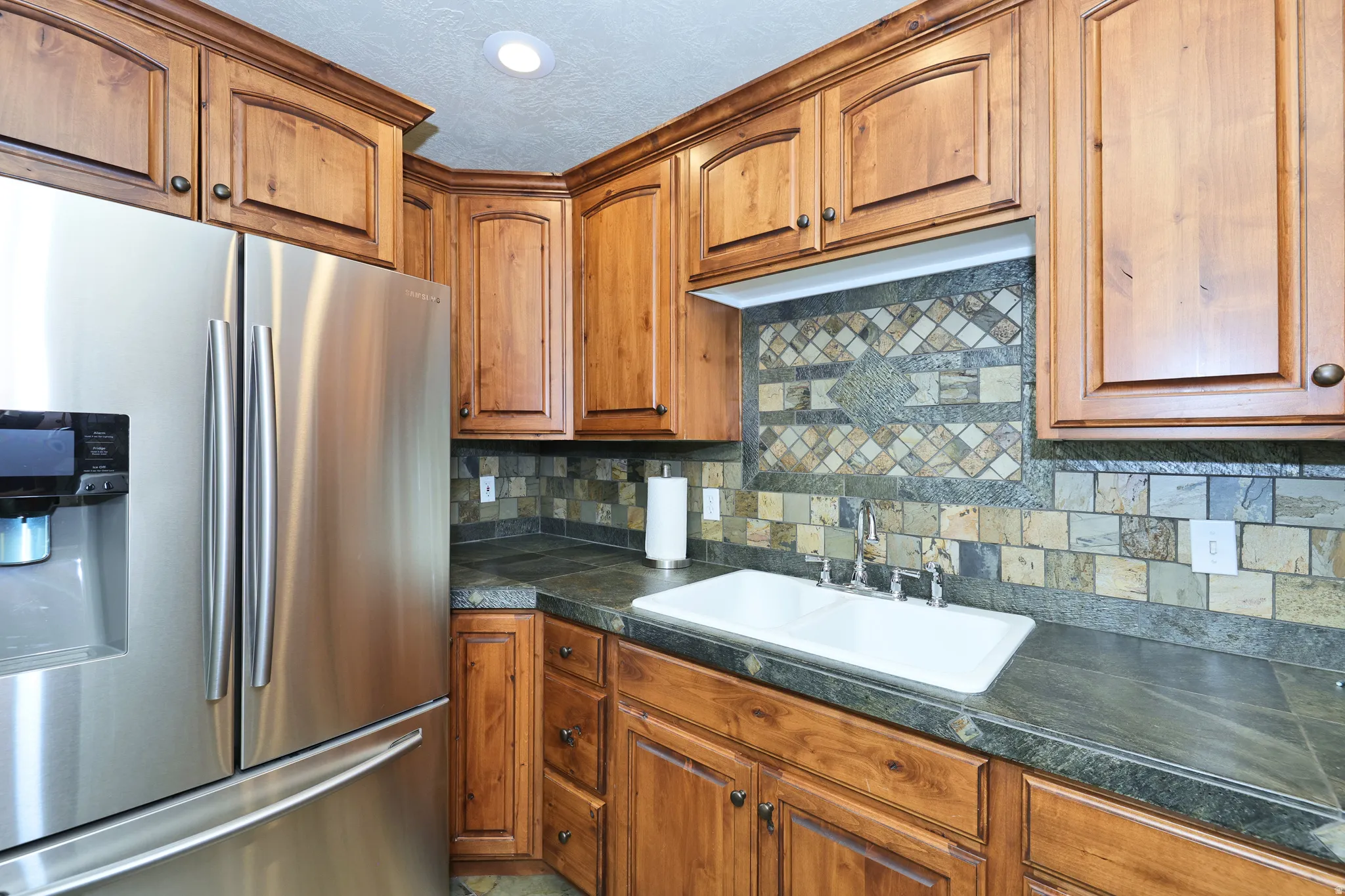Rich wood cabinetry, patterned tile backsplash, double basin sink, and integrated stainless steel refrigerator