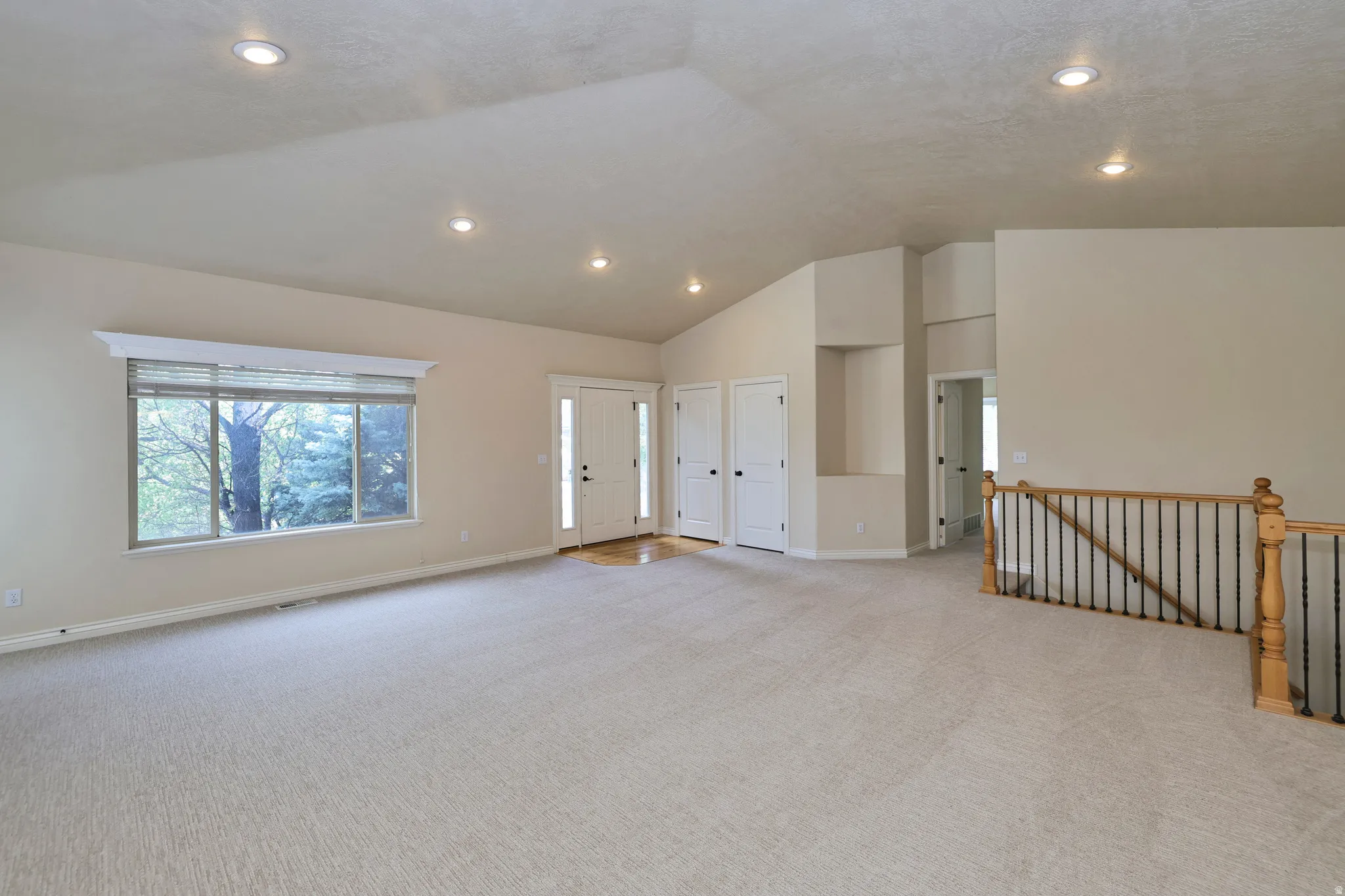 Vaulted ceiling with recessed lighting, light-toned carpeting, large window with transom, wood-finish flooring at entry, and wood and wrought iron staircase railing