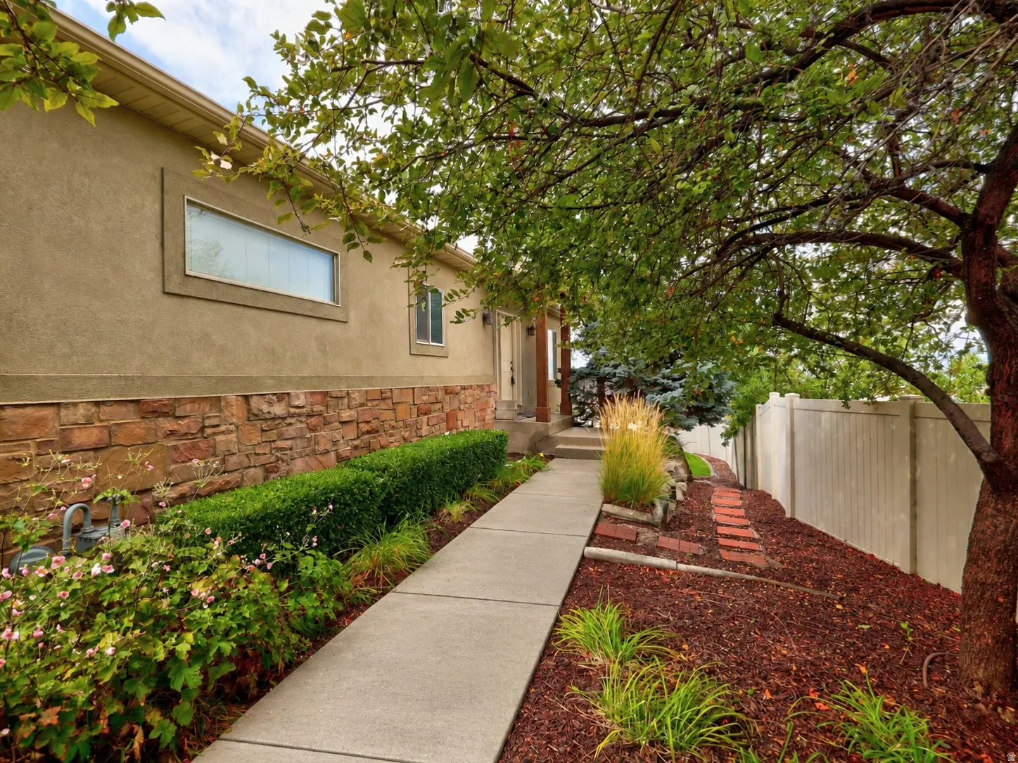 Stucco exterior with stone veneer accents, concrete pathway, established landscaping, and mature trees