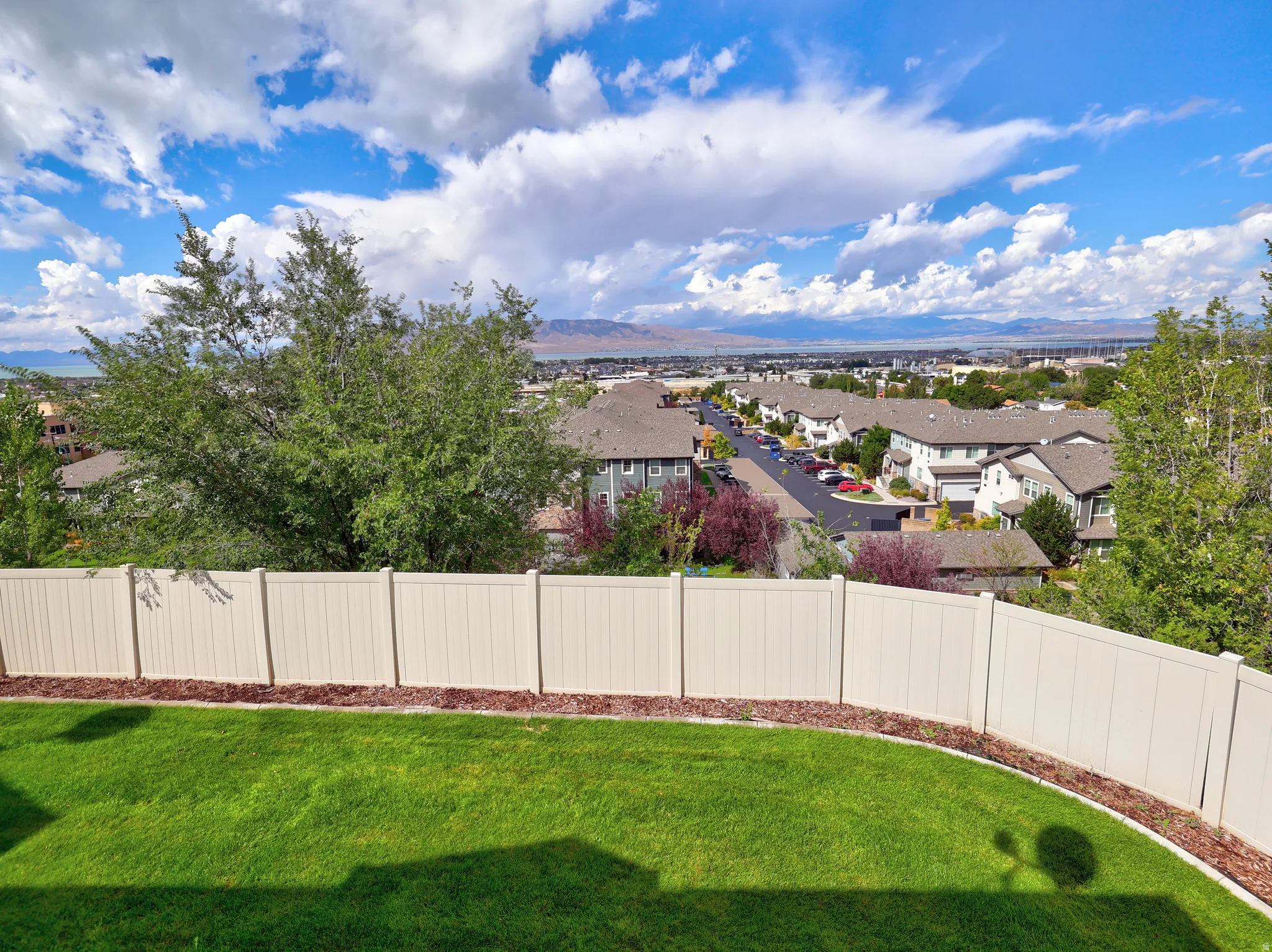 Expansive green lawn with a curved mulch border, enclosed by a perimeter fence, offering a high-elevation perspective of the surrounding residential area and distant mountains