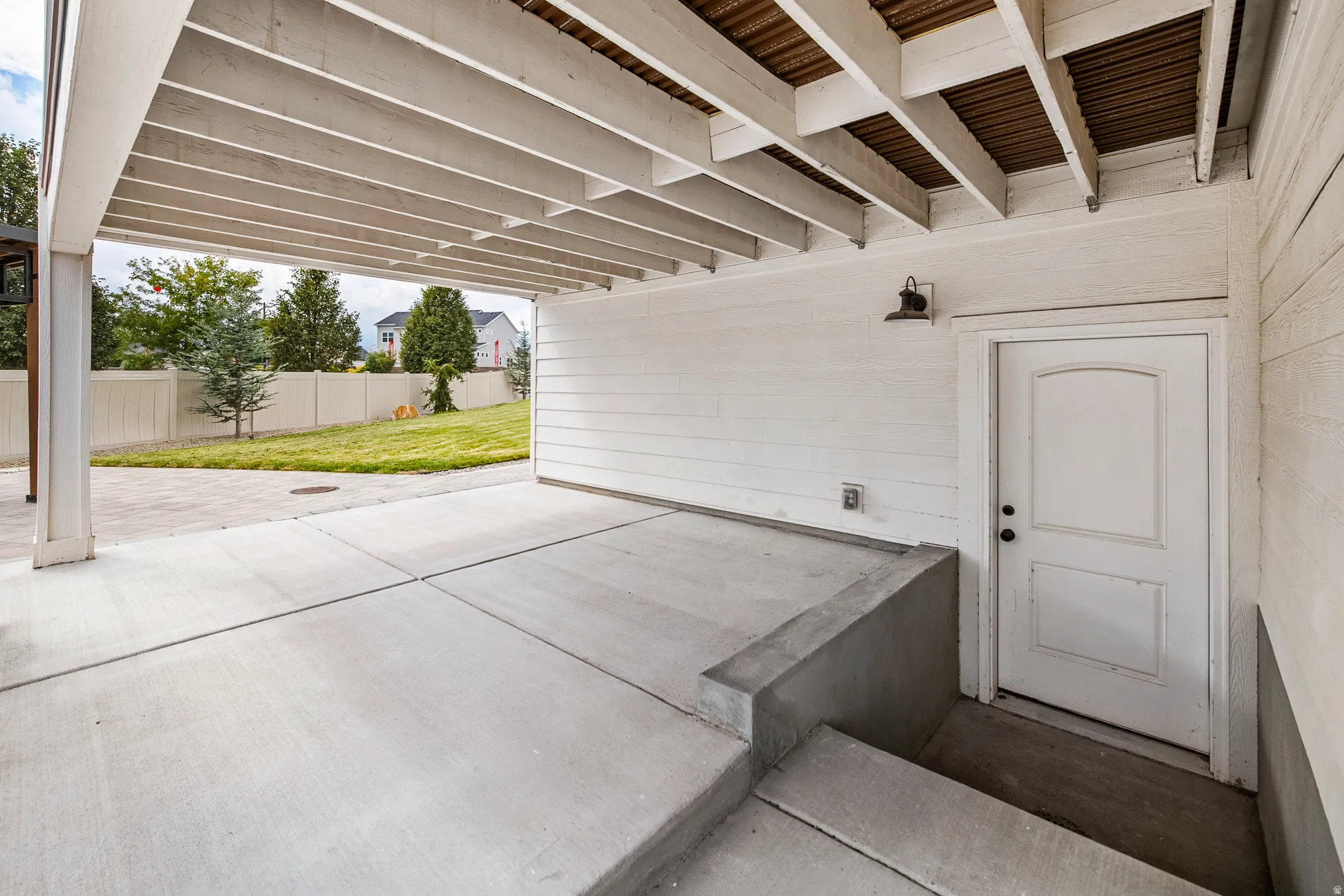 Covered concrete patio with exposed beam ceiling, white paneled siding, and an exterior door with arched paneling