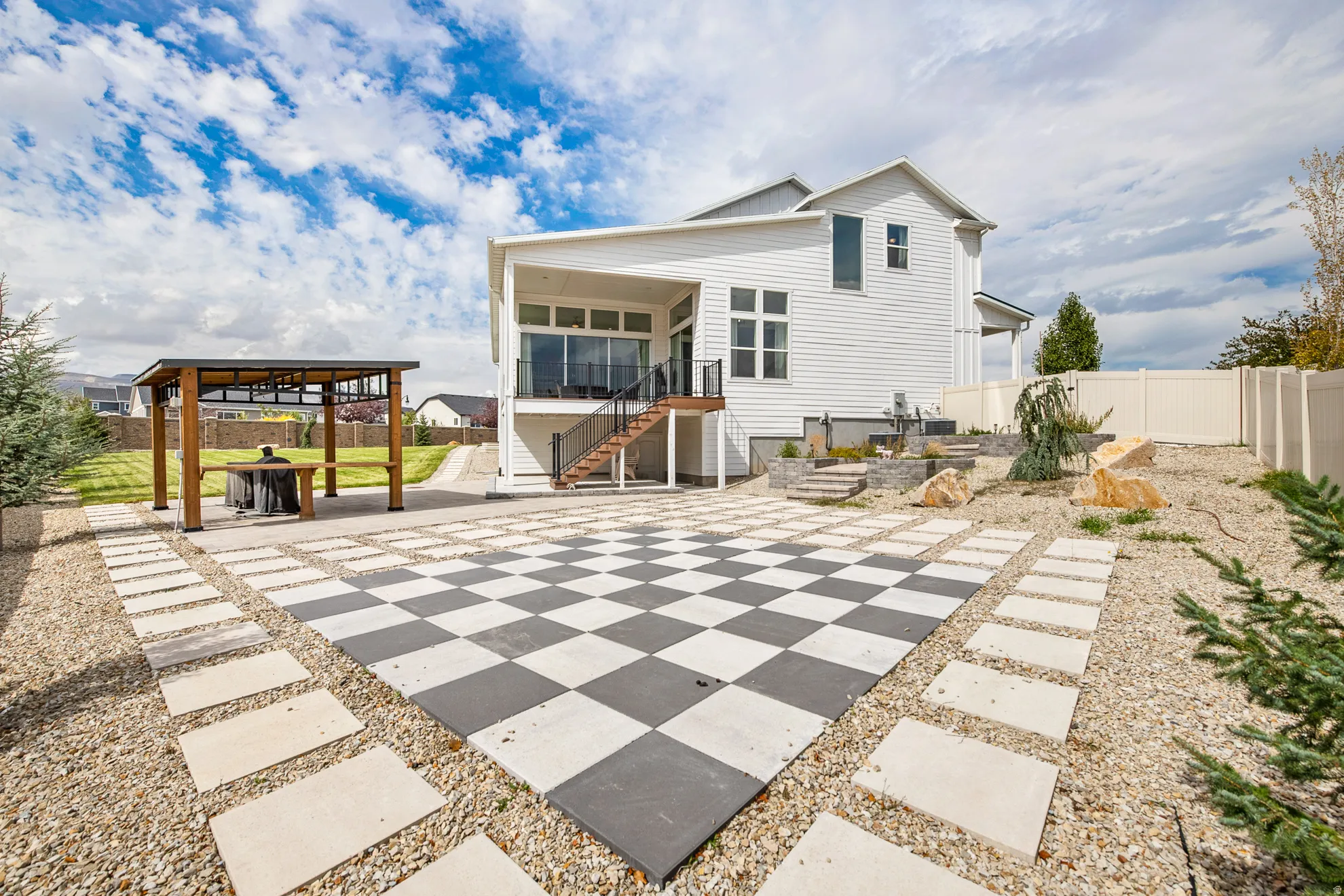 Expansive checkered patio featuring a modern pergola, a spacious deck with glass railings, and a gravel landscape with mature plantings