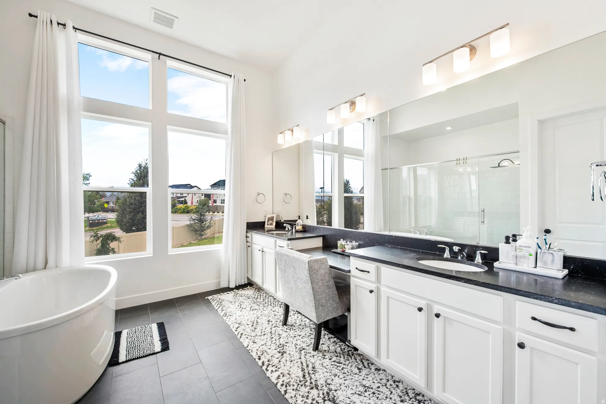 Freestanding oval soaking tub, expansive full-height windows, dual vanity with dark countertops, built-in makeup vanity, and a large mirrored wall