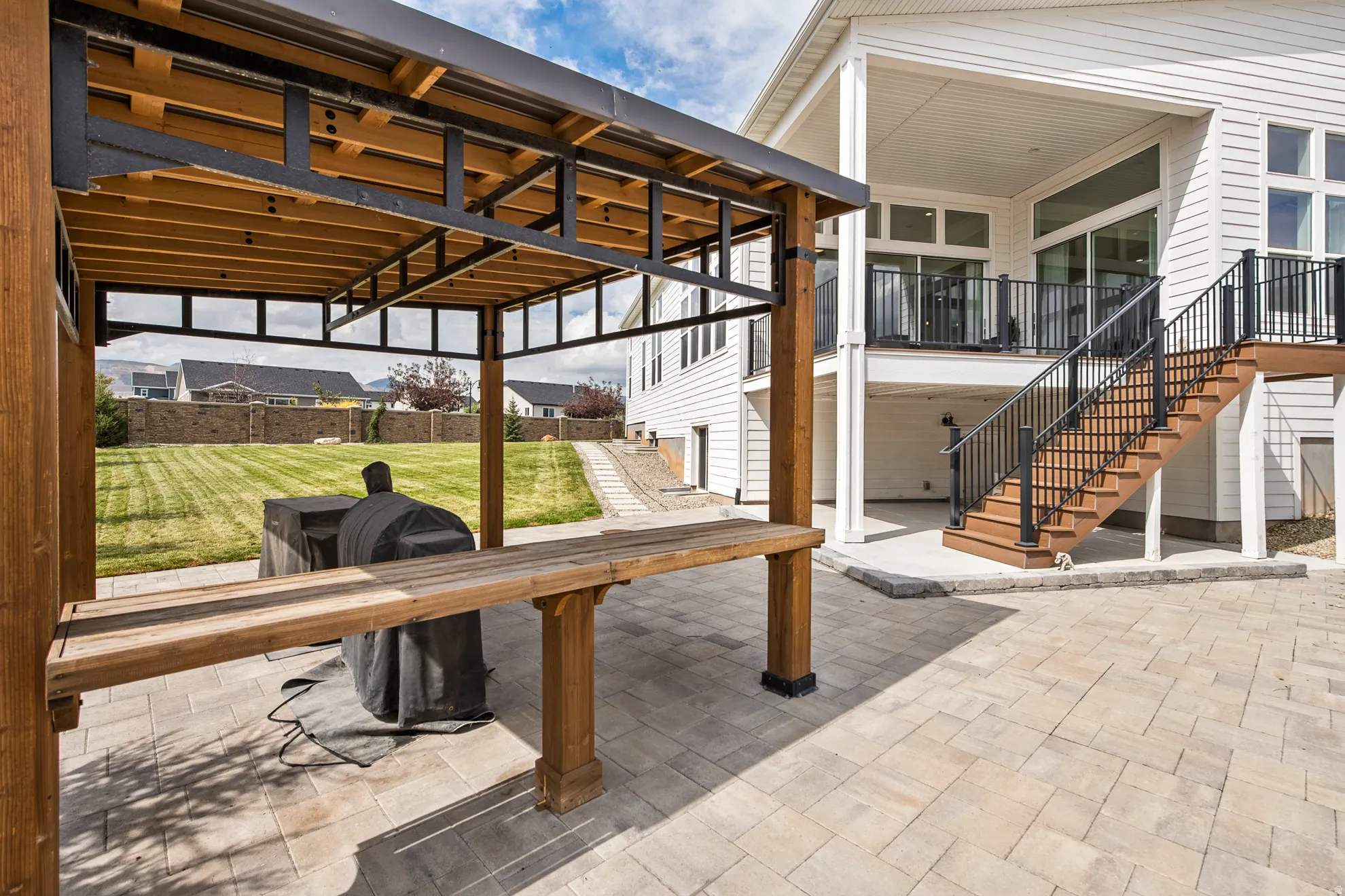 Expansive paver patio with a wood and metal pergola, a large manicured lawn, and a multi-level deck with wood-finish stairs and black railings
