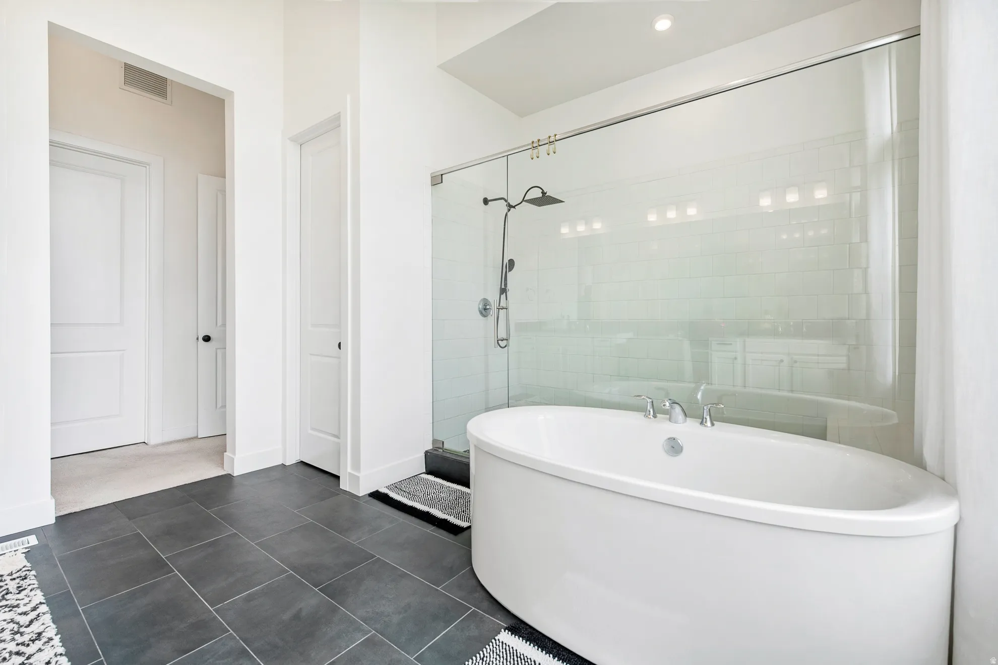 Freestanding soaking tub, glass-enclosed shower with subway tile, dark large-format floor tiles, and recessed lighting
