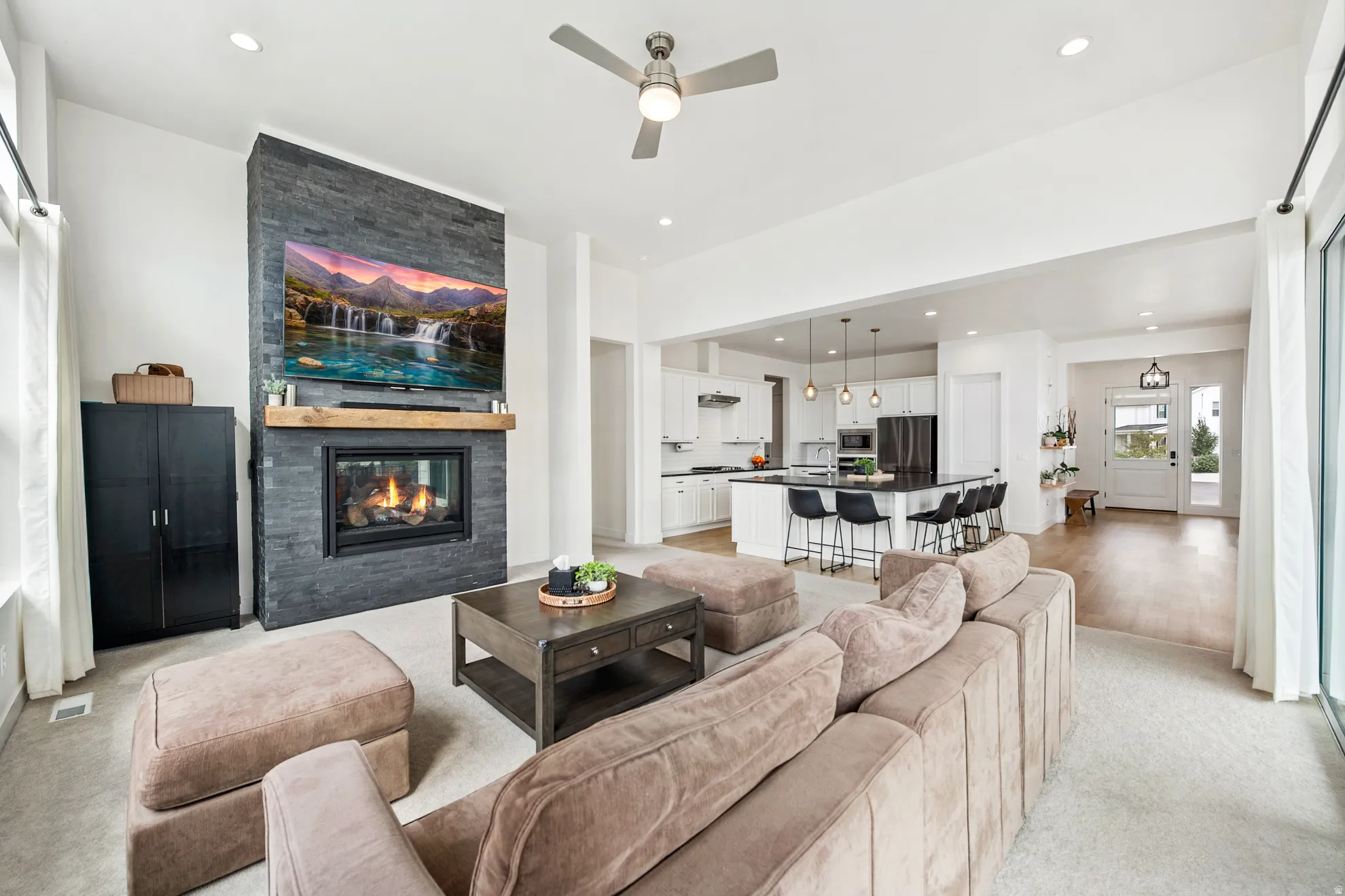 Spacious open-concept living area featuring a stacked stone fireplace with a wood mantle, recessed lighting, and a ceiling fan