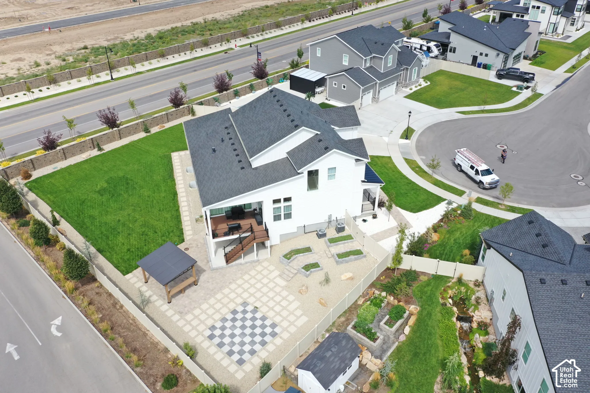 Expansive backyard featuring a large lawn area, paved patio with checkerboard design, elevated deck, and a detached pergola