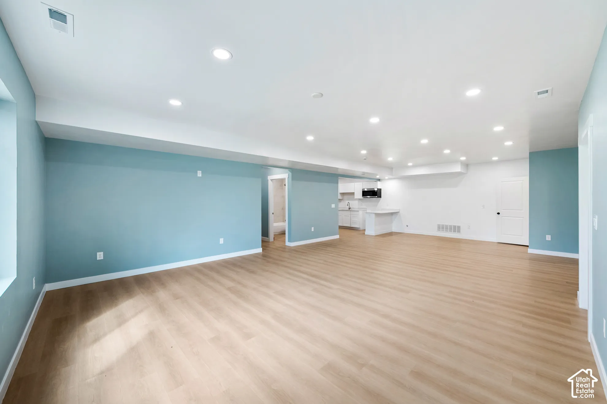 Spacious open-concept room featuring light wood-finish flooring, recessed lighting, and a partial kitchen with white cabinetry
