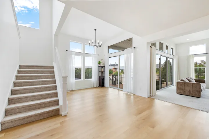 Hardwood-finish flooring throughout, complemented by a contemporary chandelier, abundant natural light from multiple windows and sliding glass doors, and a staircase with carpeted treads