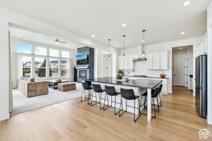 Spacious open-concept layout featuring wide plank wood-finish flooring, a large kitchen island with seating, white shaker cabinetry, a tiled backsplash, and a modern fireplace with a dark stone surround