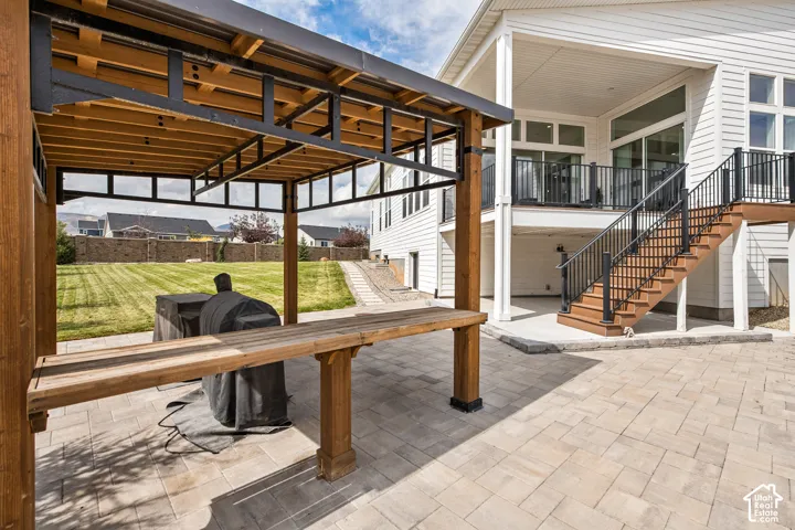 Expansive paver patio with a wood and metal pergola, a large manicured lawn, and a multi-level deck with wood-finish stairs and black railings