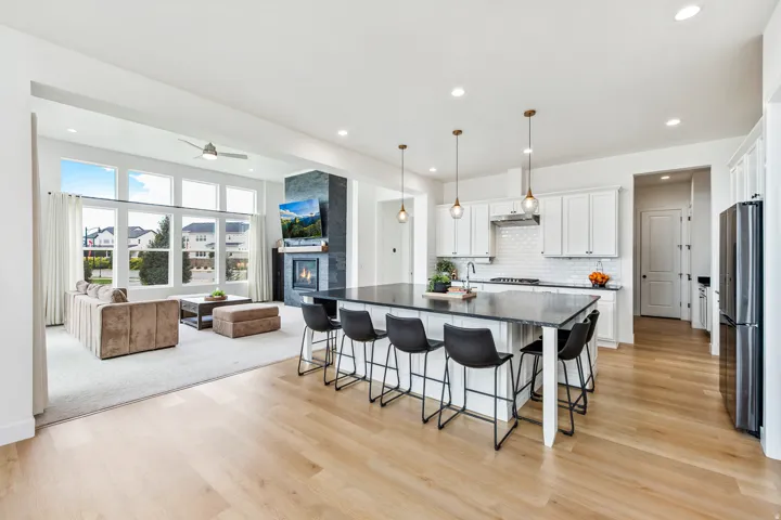 Spacious open-concept layout featuring wide plank wood-finish flooring, a large kitchen island with seating, white shaker cabinetry, a tiled backsplash, and a modern fireplace with a dark stone surround