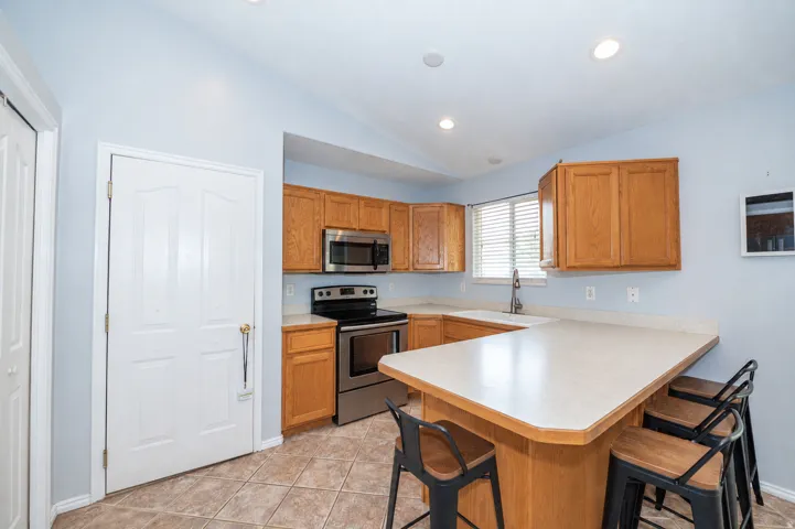 Kitchen featuring wood cabinetry, stainless steel appliances, a breakfast bar, recessed lighting, and tiled flooring. Window sink looking out to your backyard. Door entering the garage.
