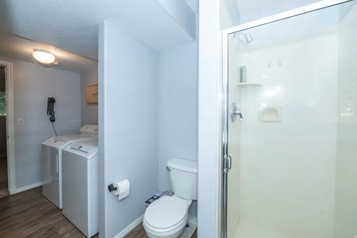 Enclosed shower with chrome fixtures, toilet, and adjacent laundry area with wood-finish flooring