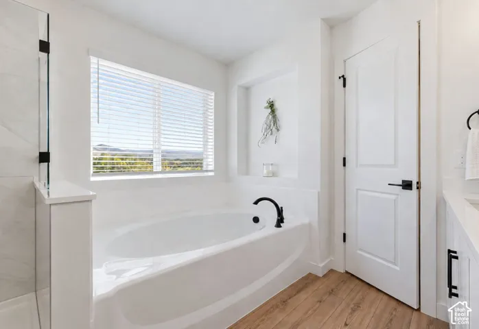 Soaking tub with black fixtures, large window with blinds, wood-finish flooring, and white paneled door