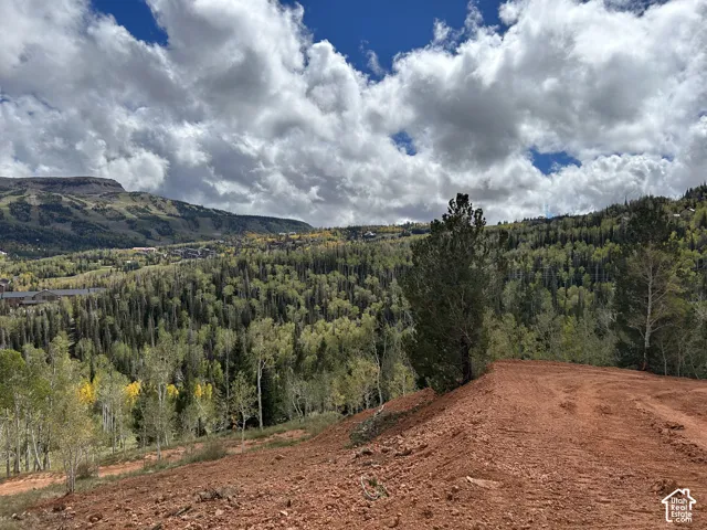 Mountainous terrain featuring a cleared building site with red soil, extensive tree coverage, and distant structures