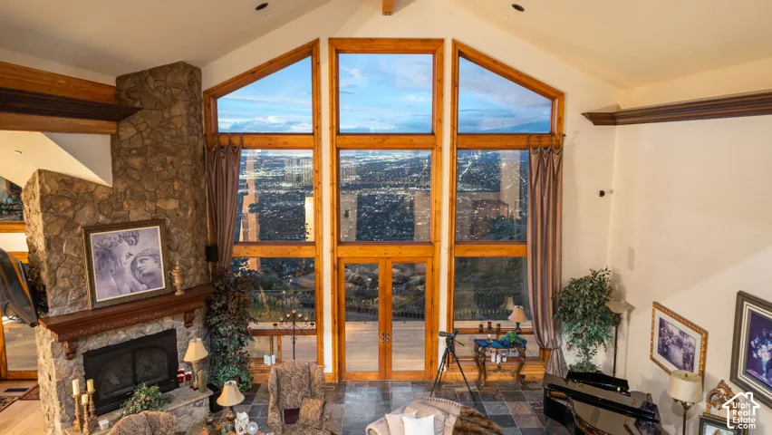 Vaulted ceiling with exposed wood beams, floor-to-ceiling windows, stone-clad fireplace with wood mantel, and stone flooring