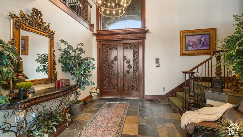 Grand foyer showcasing ornate gilded mirrors, decorative carved wooden doors with intricate paneling, multi-toned stone flooring in geometric patterns, abundant natural light from transom windows, and lush indoor greenery throughout the space