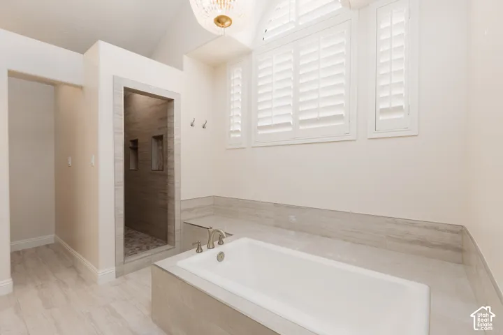 Freestanding soaking tub with brushed nickel fixtures, walk-in shower with tile surround and built-in niches, vaulted ceiling with chandelier, and plantation shutters on arched and rectangular windows