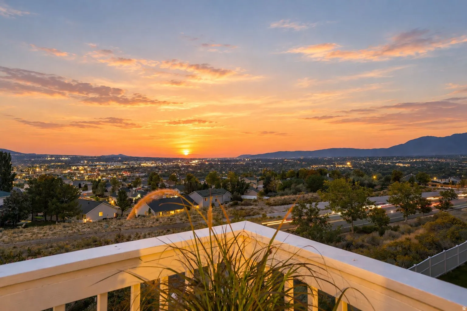 Expansive city and mountain scenery with a vibrant sunset backdrop, visible from a property featuring a white painted railing