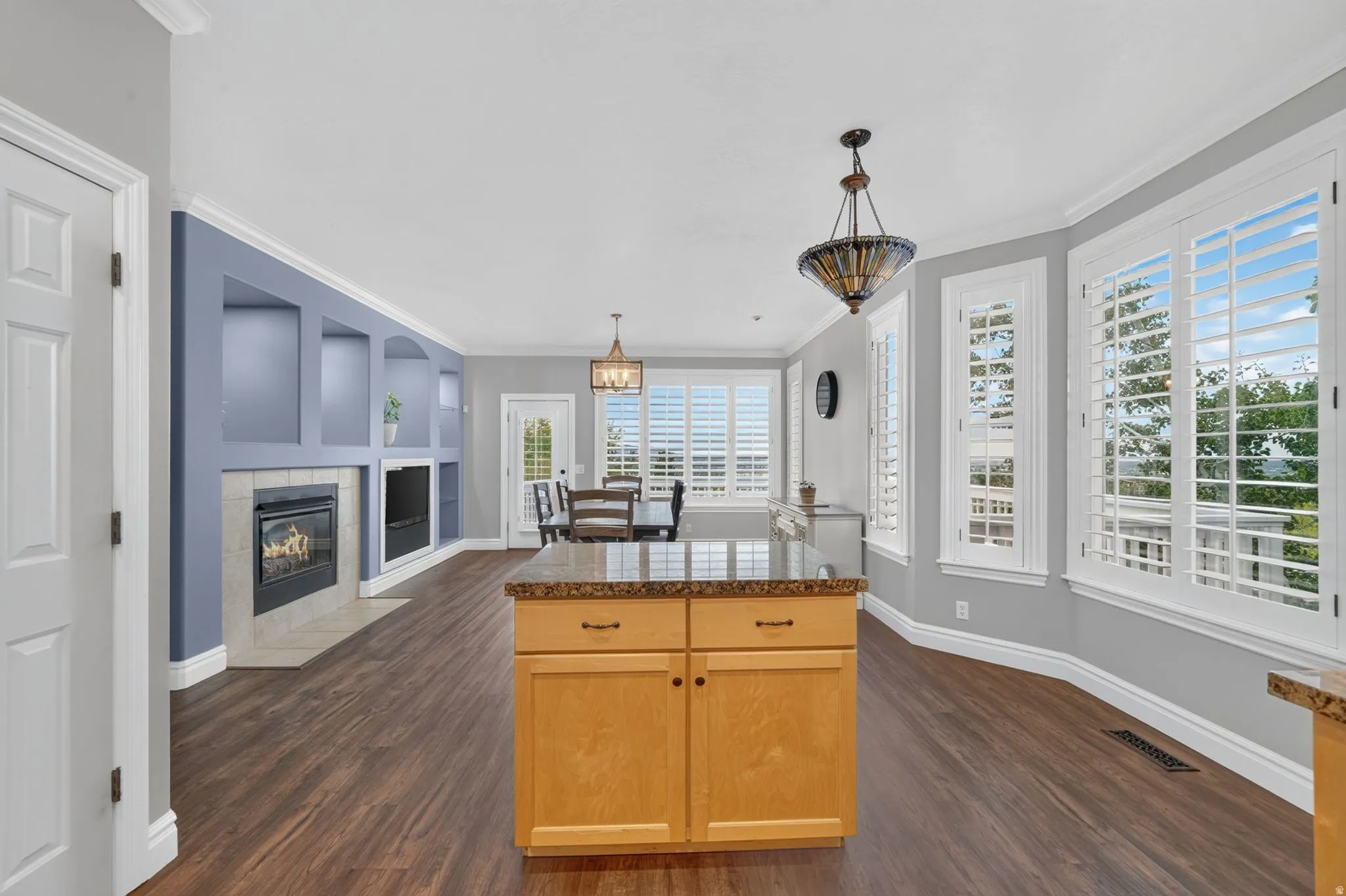 Built-in shelving with an integrated fireplace, wood-finish flooring, a kitchen island with stone countertop, plantation shutters, and decorative pendant lighting