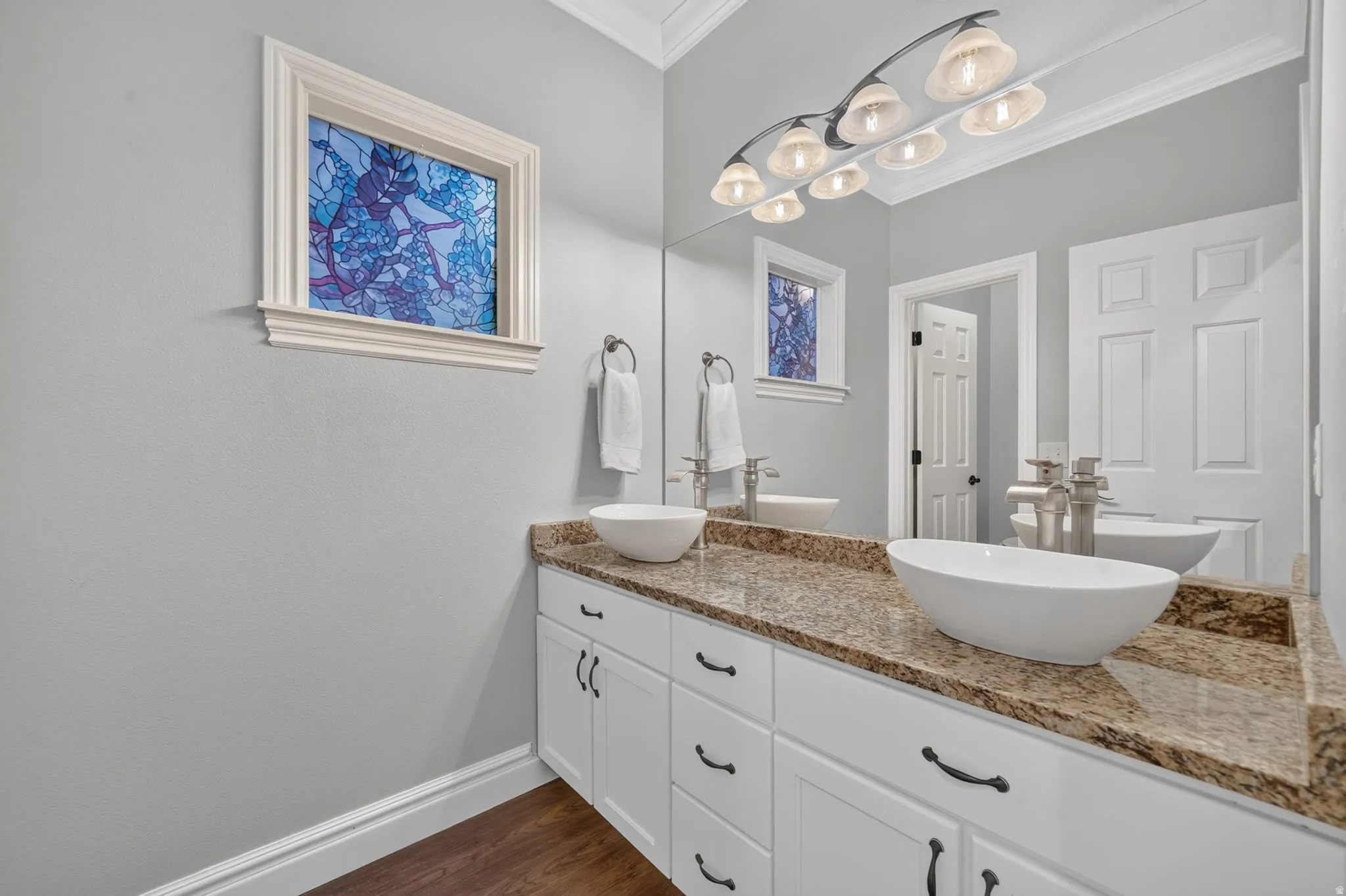 Dual vessel sinks, granite-finish countertop, white cabinetry with dark hardware, wood-finish flooring, and crown molding