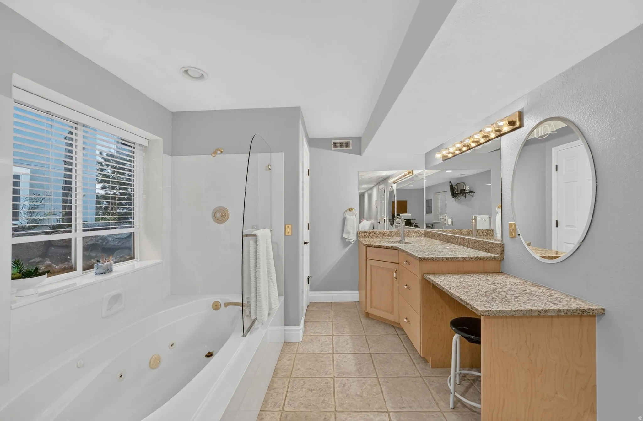 Primary bathroom featuring dual wood-finish vanities with granite countertops, an oval framed mirror with integrated lighting, a soaking tub with glass enclosure, and tile flooring throughout