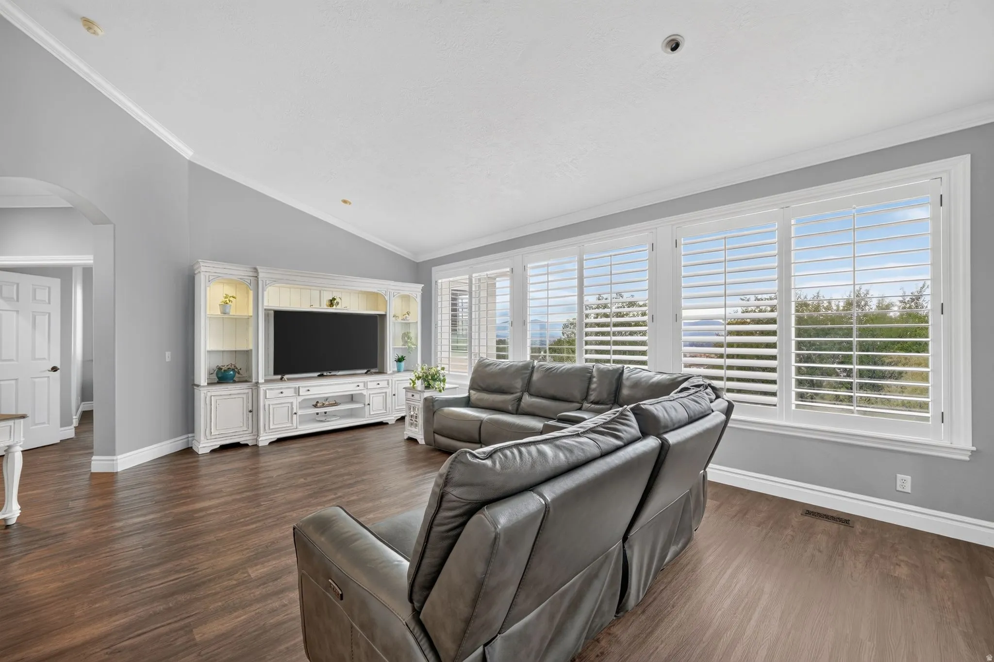 Wood-finish flooring, white built-in entertainment center, vaulted ceilings, white crown molding, and expansive windows with custom plantation shutters