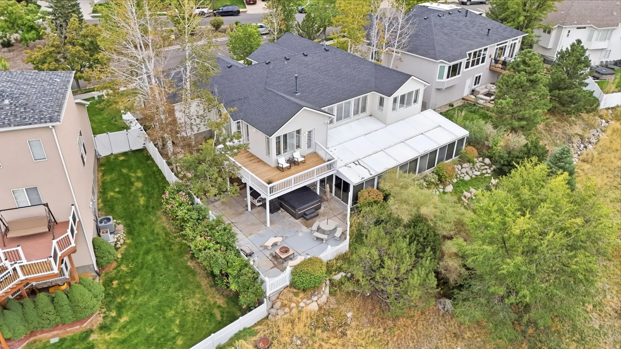 Rear exterior featuring an elevated wood-finish deck, a covered sunroom, a lower patio with a built-in fire pit, and mature landscaping