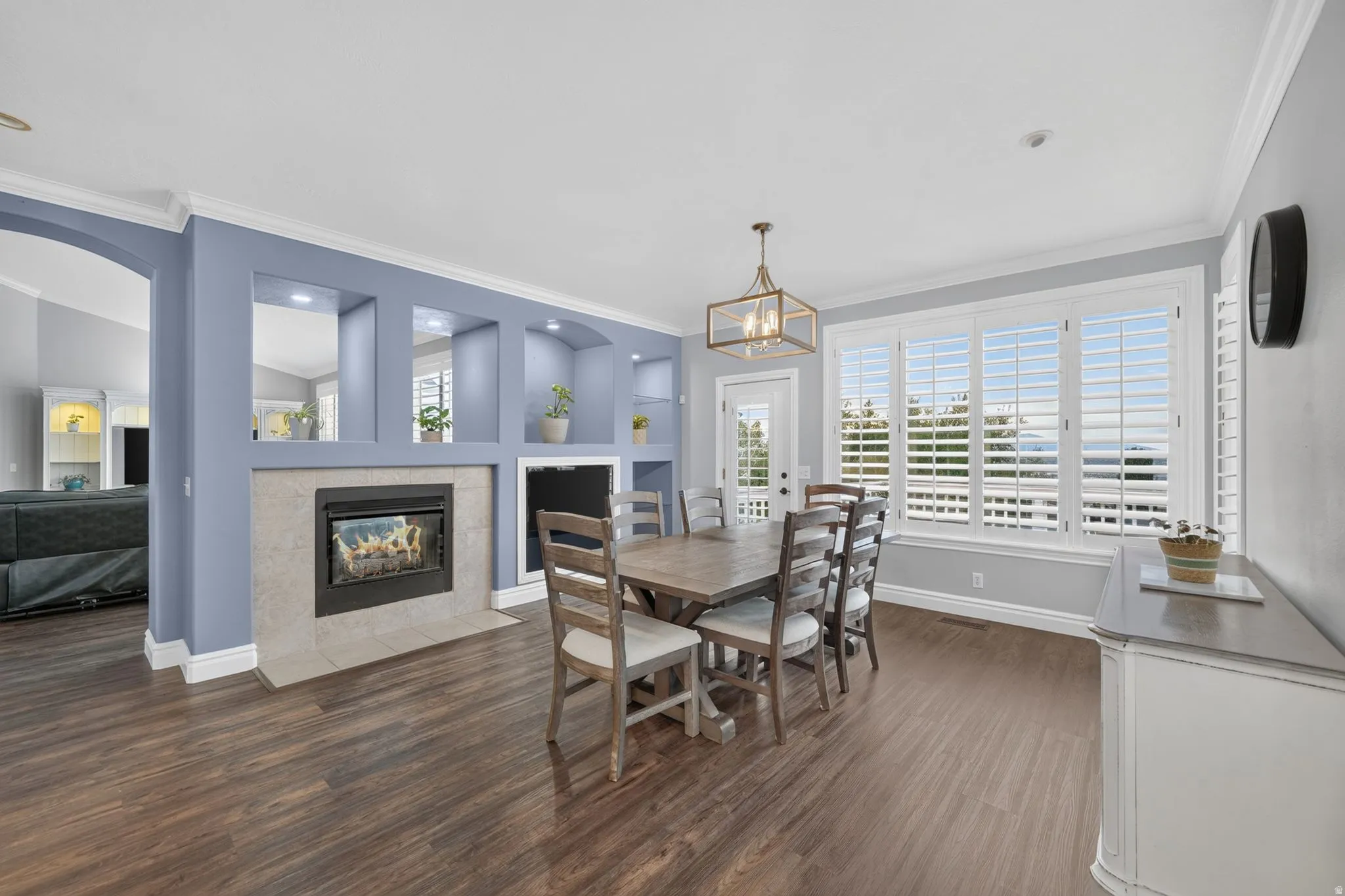 Wood-finish flooring throughout, fireplace with a tiled hearth, built-in recessed shelving, large window with plantation shutters, and crown molding