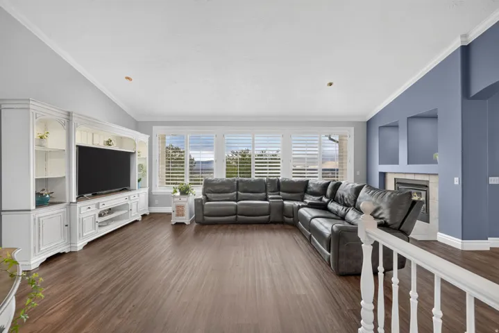 Vaulted ceiling with crown molding, wood-finish flooring, built-in entertainment center, fireplace with tile surround, and window with plantation shutters