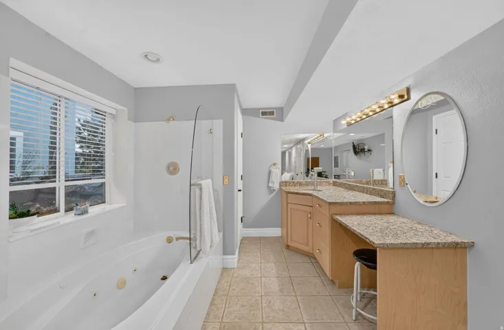 Primary bathroom featuring dual wood-finish vanities with granite countertops, an oval framed mirror with integrated lighting, a soaking tub with glass enclosure, and tile flooring throughout