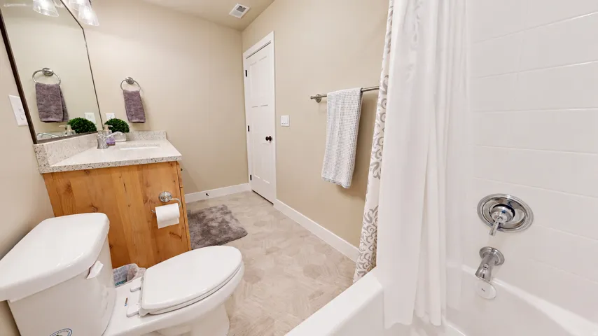 White subway tile shower surround with chrome fixtures, a wood-finish vanity with a light-toned countertop, and patterned floor tile