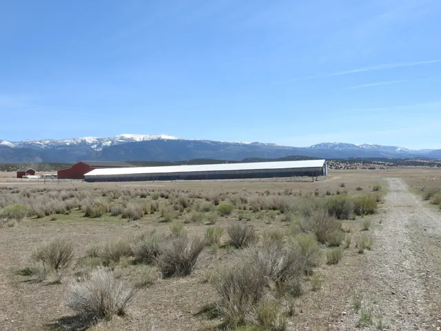 Expansive rural landscape featuring a long agricultural building with a white roof, a red barn, and a dirt road
